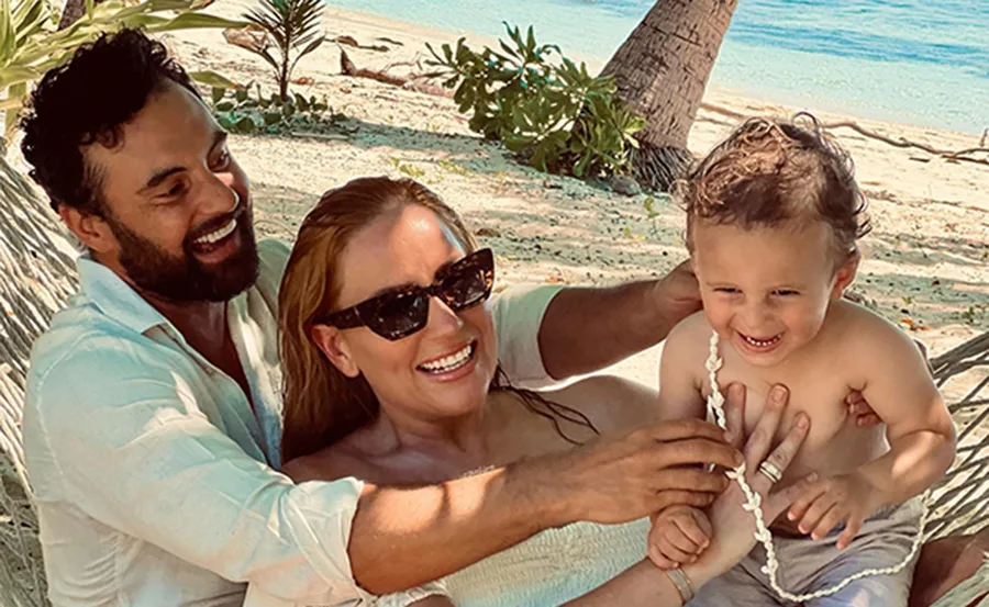 Family on a beach, smiling and playing with their toddler, surrounded by hammocks and palm trees.