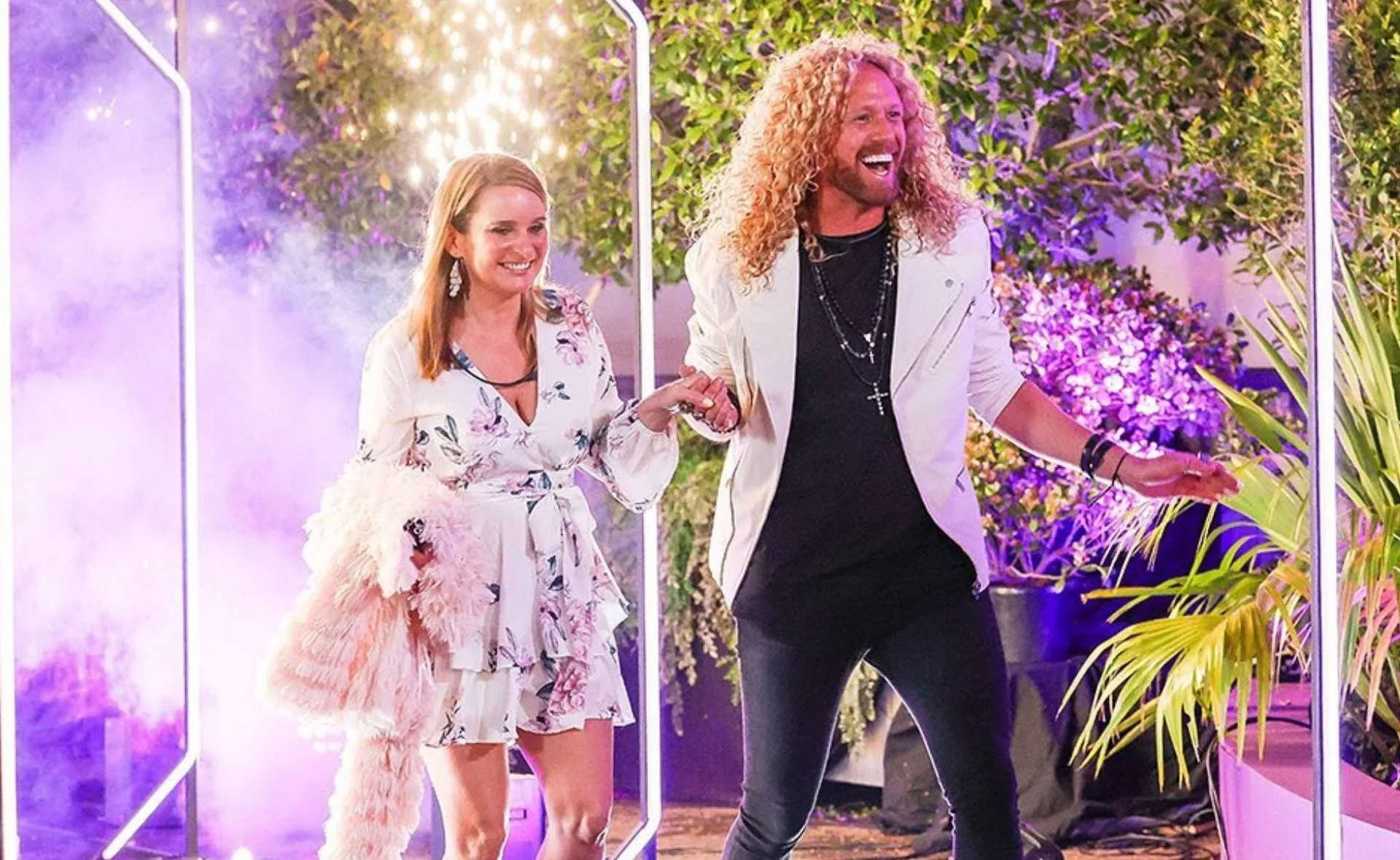 Two people in stylish outfits smiling and holding hands, walking through a brightly lit area with colorful lights and greenery.
