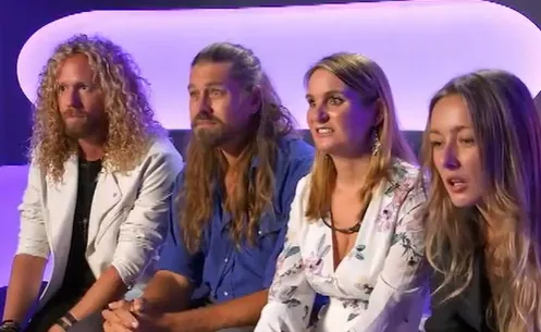 Contestants from Big Brother 2022 sit together on a couch, looking towards the camera with attentive expressions.