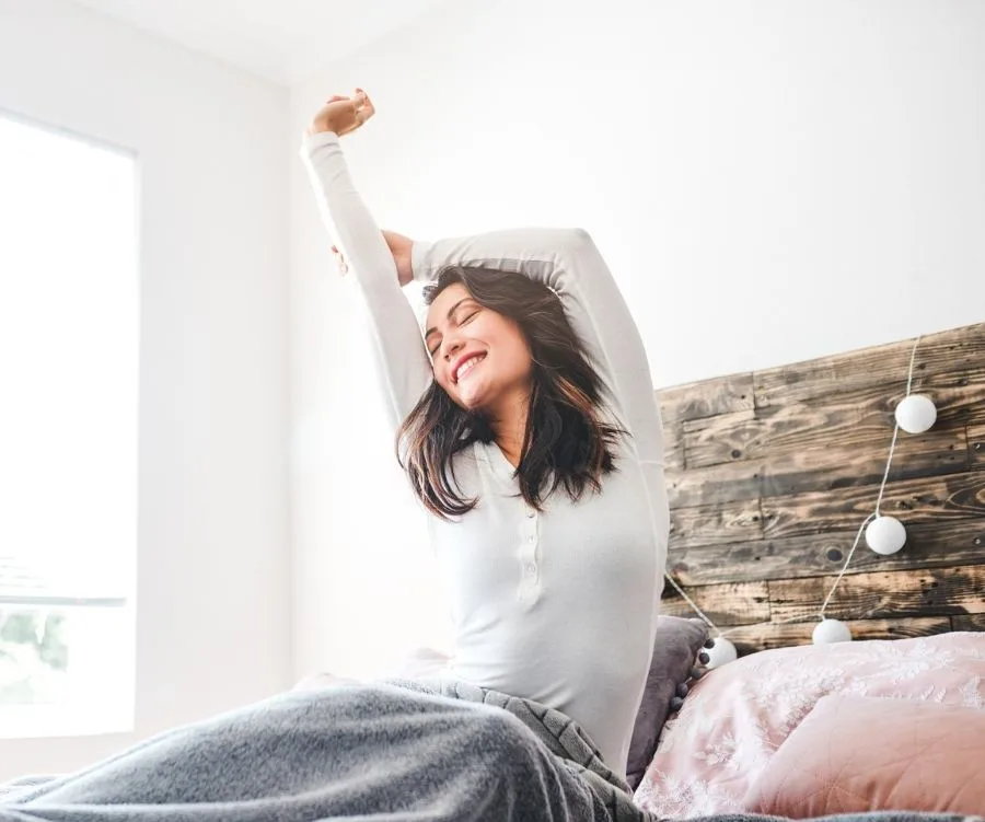 A woman happily stretches in bed with gray blanket and wooden headboard, bright room.