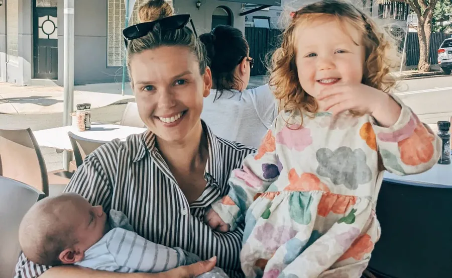 Woman smiling with a baby in her arms and a toddler in a colorful dress beside her at an outdoor café.