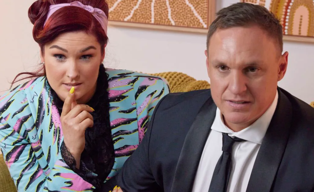 A woman with colorful outfit and man in suit sitting on couch, both appear focused.