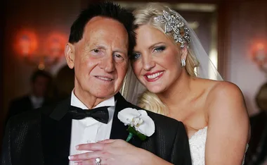 Bride in white gown and jeweled headpiece smiles, embracing an elderly man in a tuxedo with a white flower boutonniere.