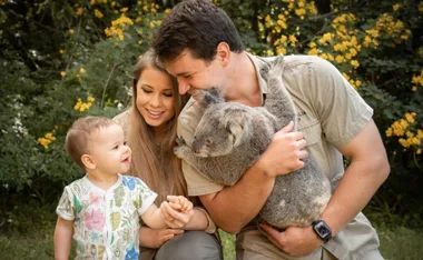 A family in a garden with a toddler holding hands, a man holding a koala, and yellow flowers in the background.