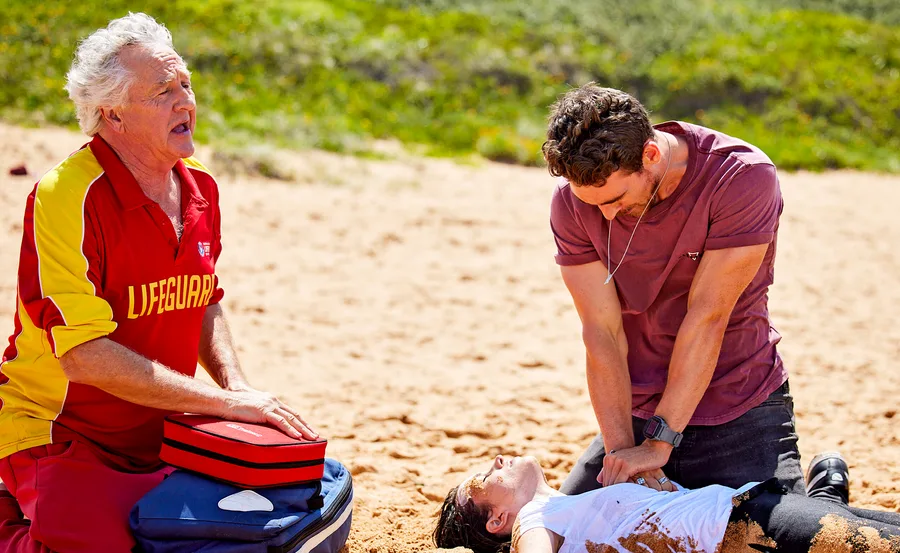 Man performing CPR on a beach, assisted by lifeguard in red and yellow uniform with medical kit nearby.