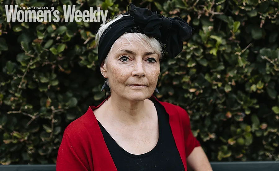A woman in a red cardigan and black headscarf, standing in front of green foliage.
