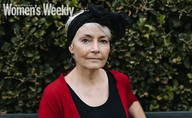 A woman in a red cardigan and black headscarf, standing in front of green foliage.