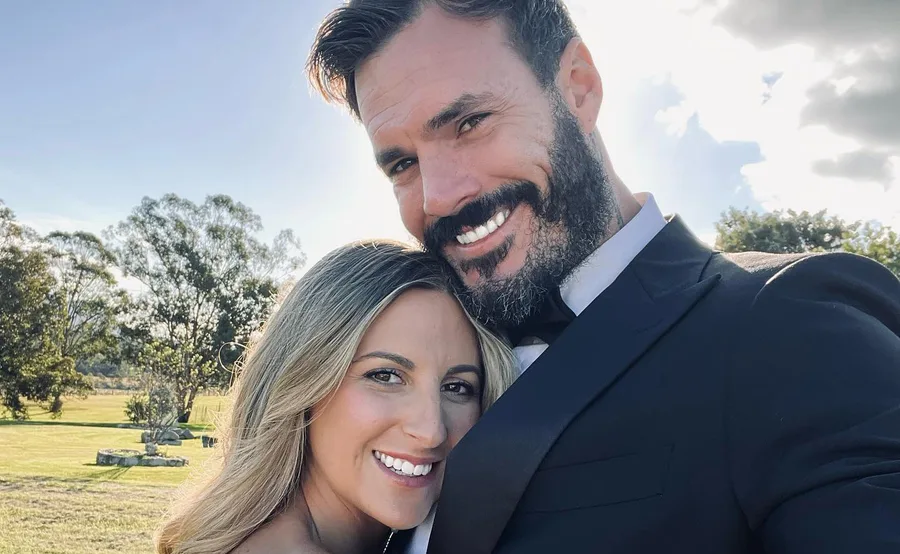 Couple smiling outdoors, man in suit and woman with long hair, with trees and blue sky in the background.