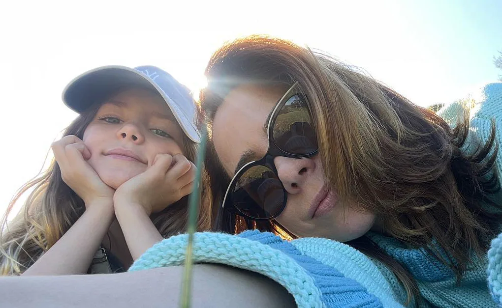 Mother and daughter lying on grass; daughter in a blue cap, mother in sunglasses, both smiling slightly.