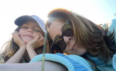 Mother and daughter lying on grass; daughter in a blue cap, mother in sunglasses, both smiling slightly.