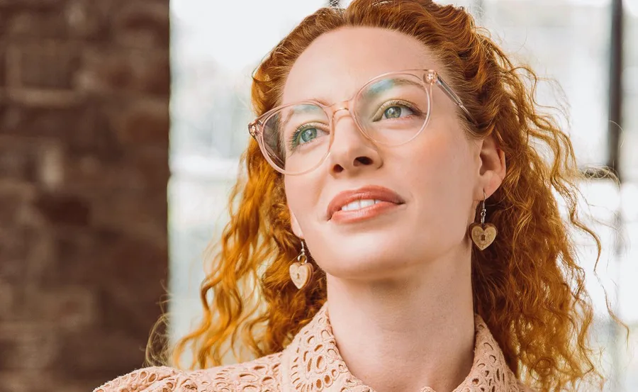 Female with red curly hair, glasses, and heart earrings, wearing a lace top, looking up thoughtfully indoors.