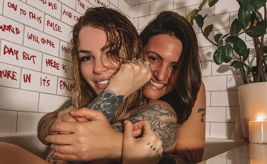 Two people with tattoos smiling in a bathtub, with a white tiled wall and a potted plant in the background.