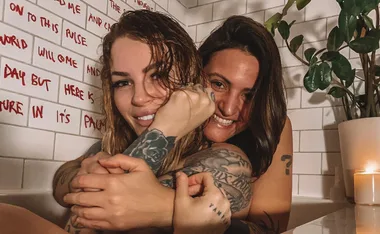 Two people with tattoos smiling in a bathtub, with a white tiled wall and a potted plant in the background.