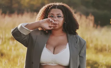 Woman in white lace bra with open blazer, standing in field, hand near face, curly hair, glasses, warm lighting.