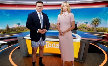Two TV hosts on set; one in a suit jacket and boxers, the other in a pink dress, standing by a news desk.