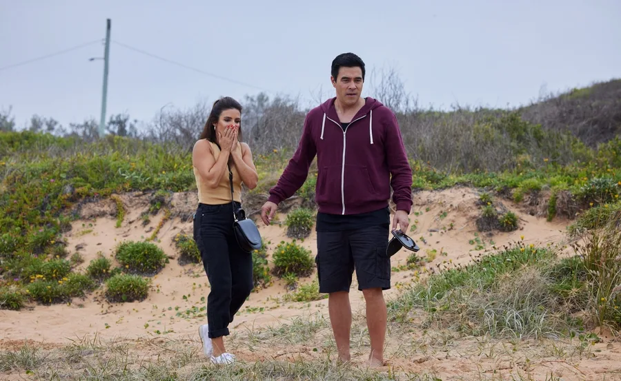 Two people, a woman looking shocked and a man holding shoes, standing on a sandy beach with grass in the background.