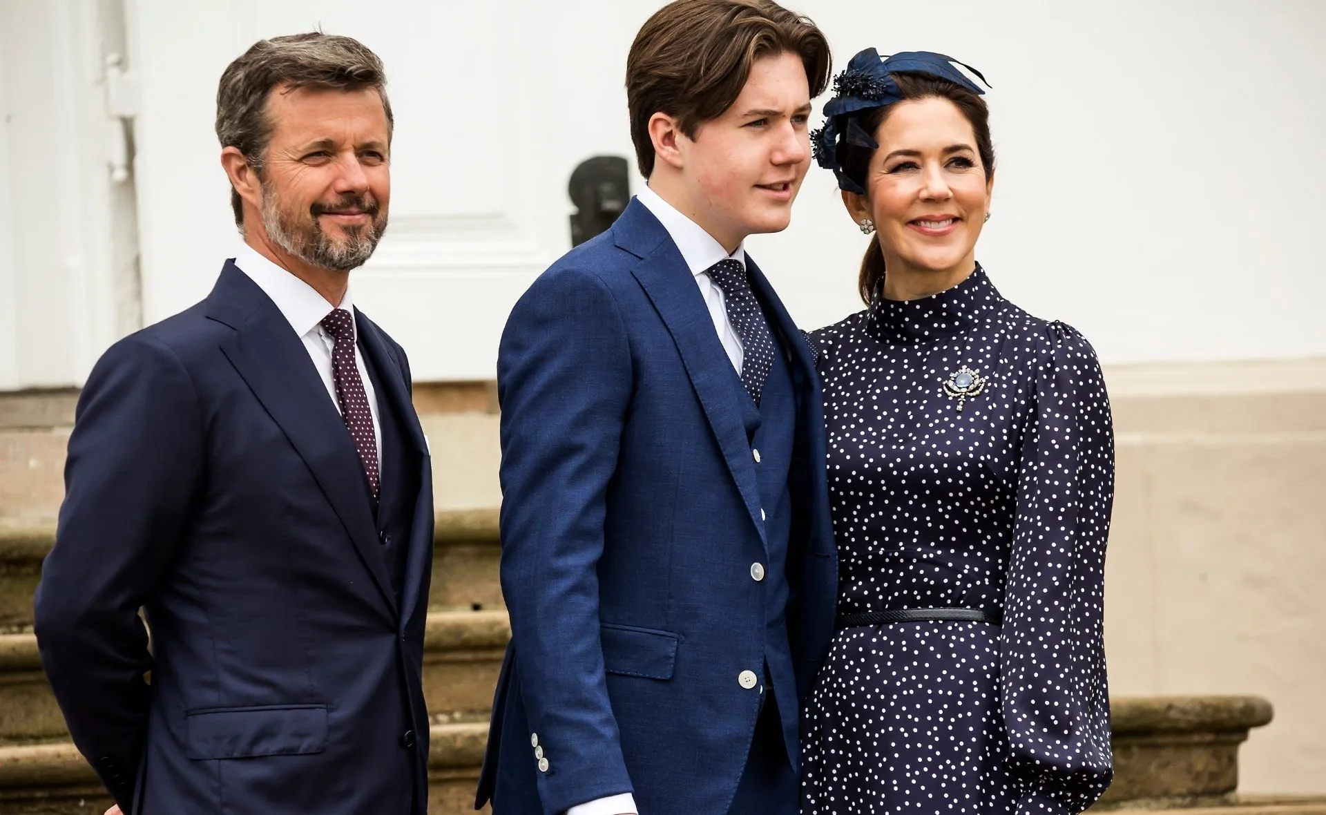 Royal family members smiling, all dressed in formal attire, stand outside near stairs.