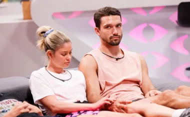 Two contestants from Big Brother Australia 2022, one with a pink shirt, sit closely on a couch, focused and pensive.