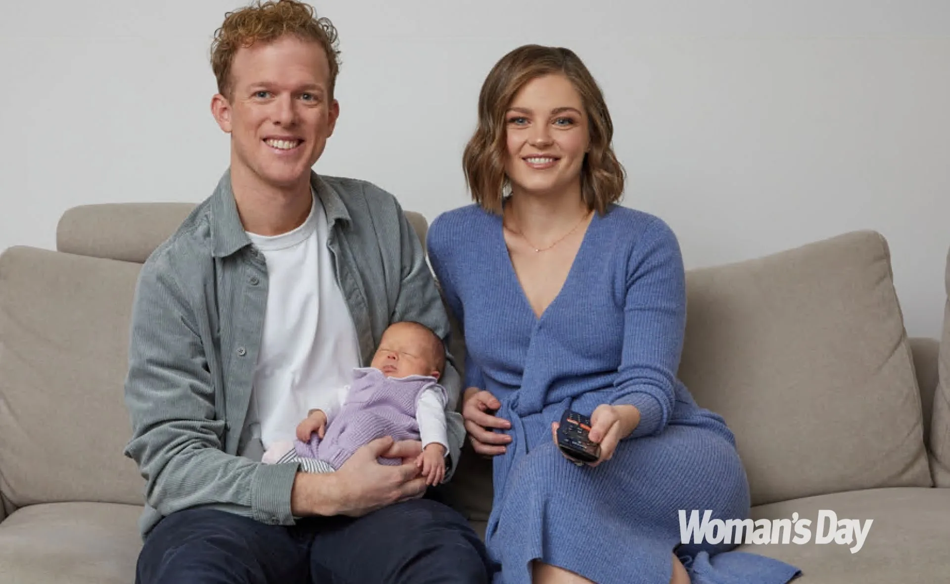 A couple smiles on a sofa, holding a newborn baby dressed in a purple outfit.