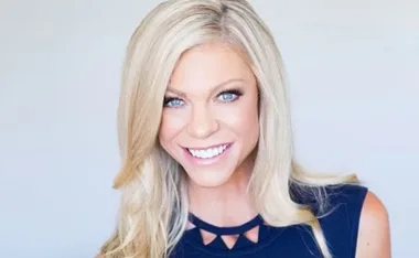 A woman with long blonde hair smiling, wearing a navy blue top with triangle cutouts.