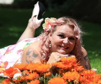 An older woman with pink hair and tattoos lies on grass, smiling among vibrant orange flowers, wearing a floral dress.