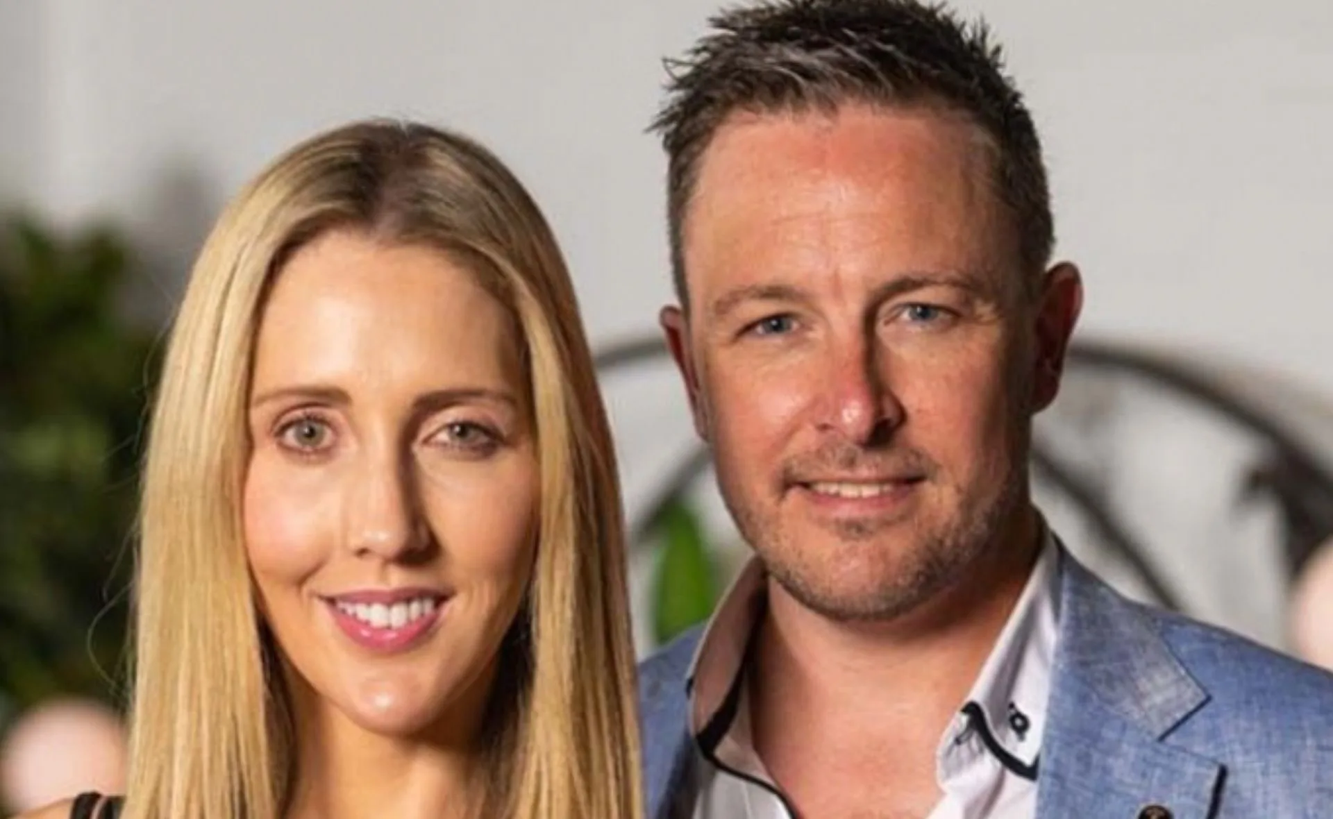 A man and woman standing together, smiling, indoors with a blurred background.