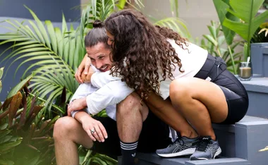 Couple sitting on stairs, woman hugging man from behind, surrounded by plants.