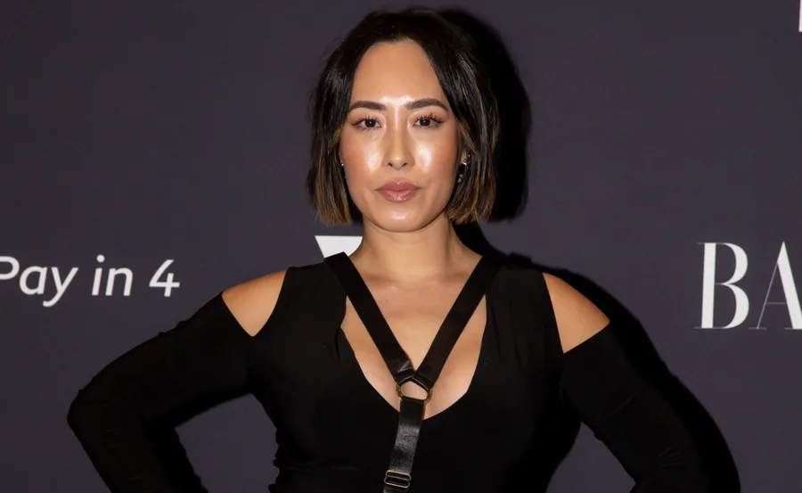 A person in a black outfit poses at an event with a dark background displaying text.