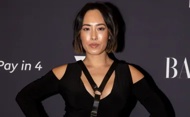 A person in a black outfit poses at an event with a dark background displaying text.
