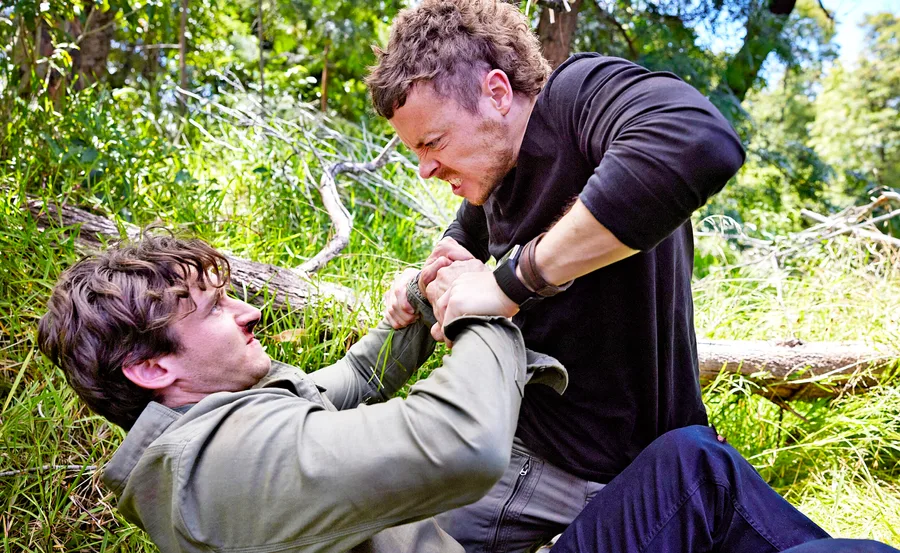 Two men struggle in a grassy area, one on top holding the other down, both showing intense expressions.