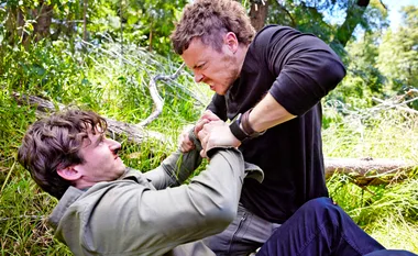 Two men struggle in a grassy area, one on top holding the other down, both showing intense expressions.