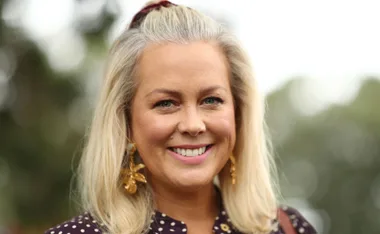 Smiling woman with blonde hair, wearing polka dot outfit and gold earrings, in an outdoor setting.