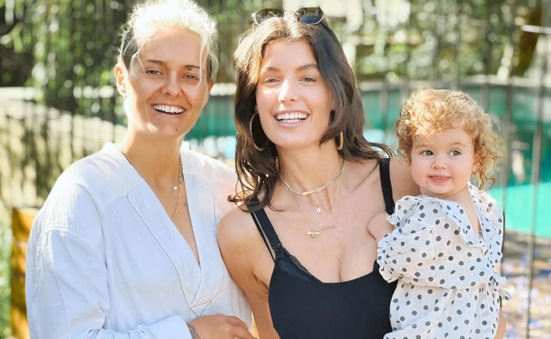 Smiling group of three, two adults and a baby in polka dot outfit, in sunny outdoor setting.