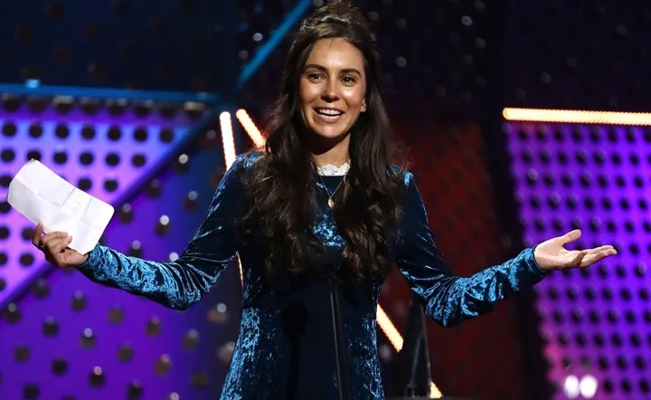 A woman in a blue dress speaks on stage, holding a paper, with a colorful, illuminated backdrop behind her.