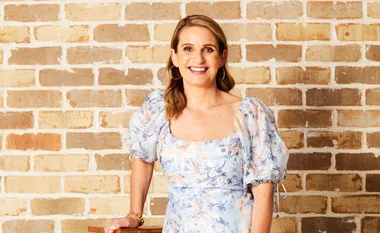 Woman smiling in floral dress, standing against a brick wall.