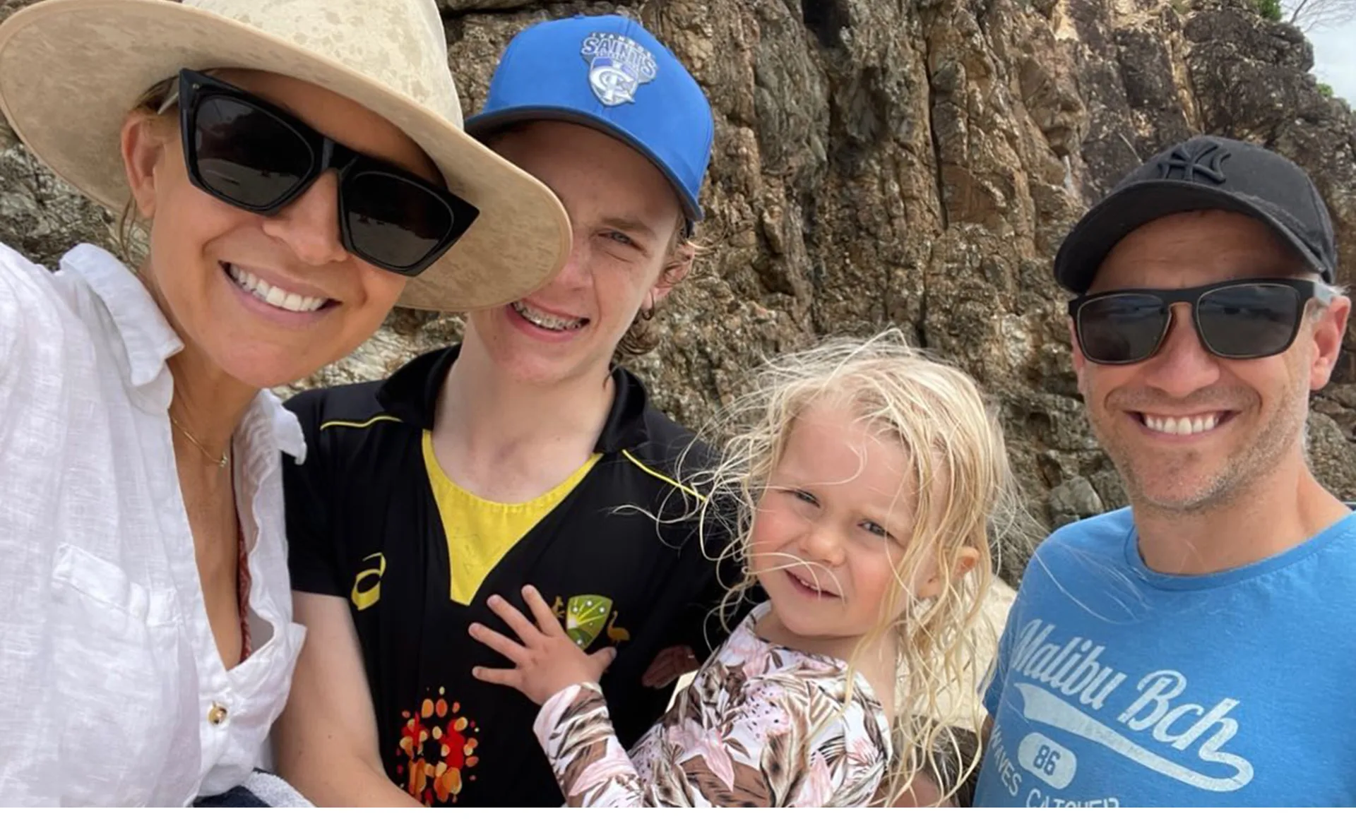 A family smiling in a selfie at a rocky beach, all wearing casual summer clothing and sunglasses.
