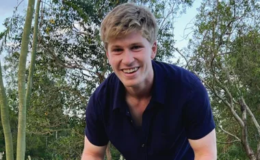 A smiling young man in a dark shirt outdoors with trees in the background.