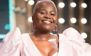Smiling individual with short hair and pink dress, soft lights in background.