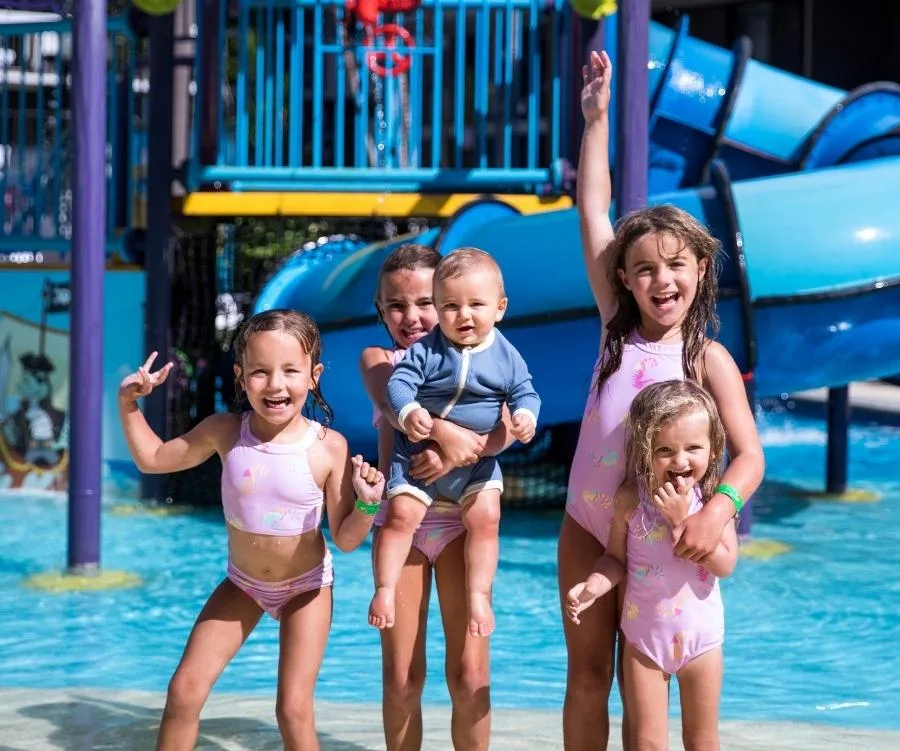 Children in swimsuits laughing and posing in a water park with slides in the background.