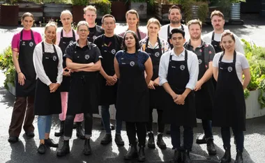 A group of MasterChef contestants wearing aprons, posing outdoors with greenery in the background.
