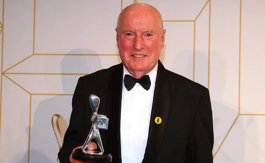 Man in a tuxedo holding a silver Logie award trophy, standing against a patterned background.