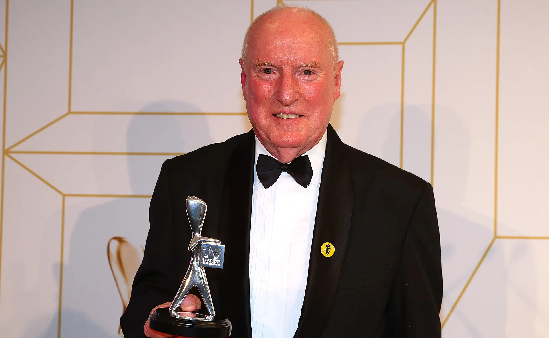 Man in a tuxedo holding a silver Logie award trophy, standing against a patterned background.