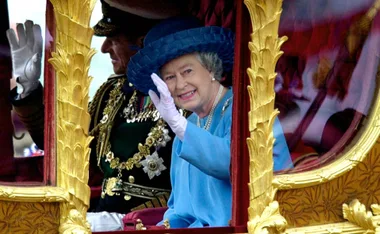 Woman in blue outfit and hat waves from a gilded carriage window.