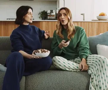 Two women sitting on a couch; one has a bowl of snacks, and the other holds a remote, both appear to be talking.