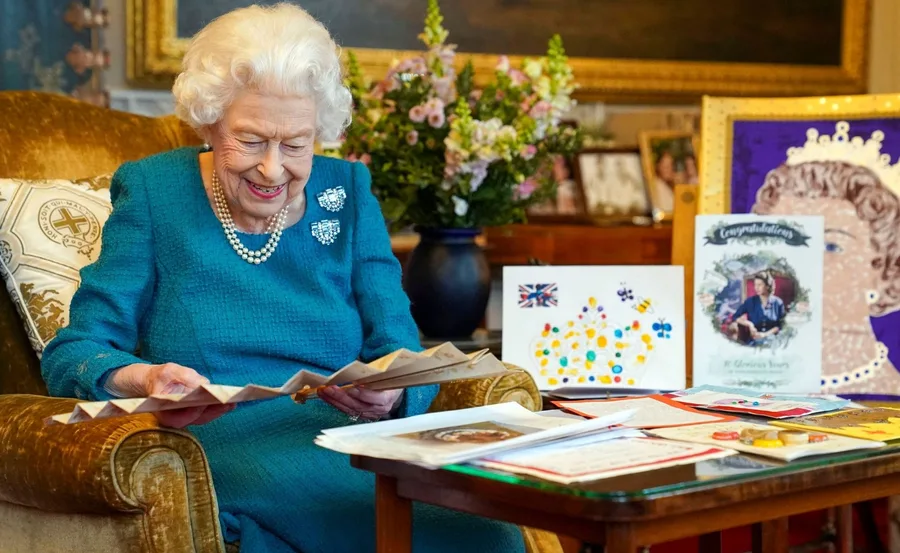 An elderly woman in a blue dress smiles while reading letters, surrounded by flowers and greeting cards on a table.