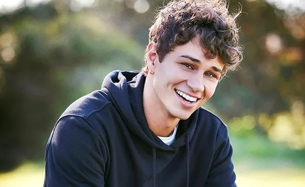 Young man with curly hair smiling outdoors, wearing a dark hoodie against a blurred green background.