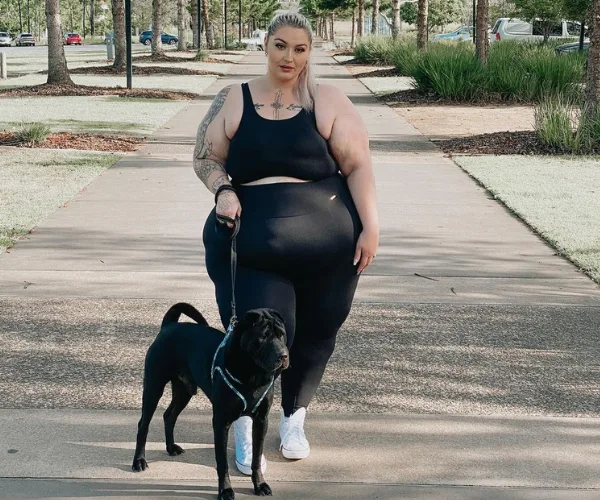 Woman in black workout clothes walking a black dog on a tree-lined path.