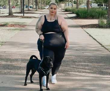 Woman in black workout clothes walking a black dog on a tree-lined path.