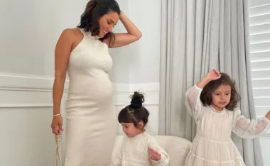 Pregnant woman in a white dress smiles with two young girls, one standing, one sitting, in a softly lit room.