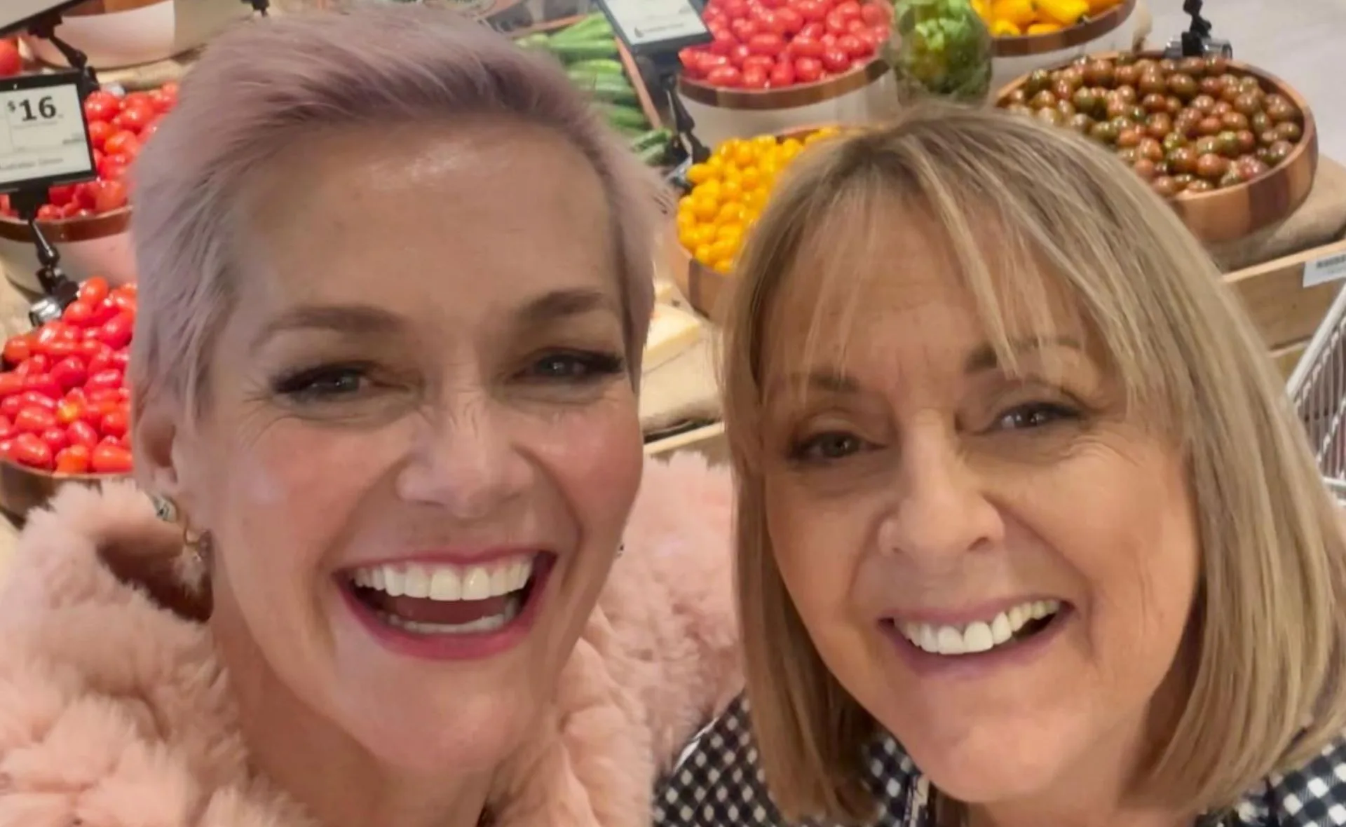 Two women smiling in front of a colorful display of tomatoes and vegetables in a grocery store.
